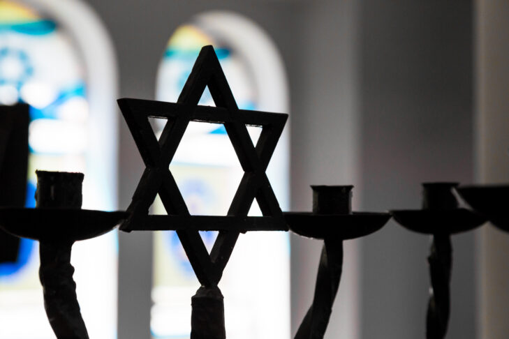 Close up image depicting the Jewish religious symbol of the star of David inside a synagogue. The star is in silhouette, while in the background stained glass windows are blurred out of focus. Horizontal colour image with copy space.
