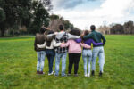 Group of six teenager friends embracing together at the park, rear view. Teamwork and cooperation concept with people together, sharing a common purpose.