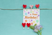 Happy birthday card with red heart clip and purple flower on blue background