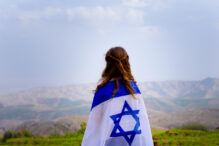 Little patriot jewish girl standing and enjoying great view on the sky, valley and mountains with the flag of Israel wrapped around her. Memorial day-Yom Hazikaron and Yom Ha'atzmaut concept.
