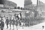 The Coliseum forms the background as a contingent of German Hitler Youth, carrying a Nazi banner, march through ancient Rome, Sept. 28, 1936. Taking advantage of the space between the standard bearer and the scouts, an Italian youngster darts across to a better vantage point. (AP Photo)
