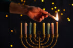 partial view of man lighting up candles in menorah on black background with bokeh lights on Hanukkah