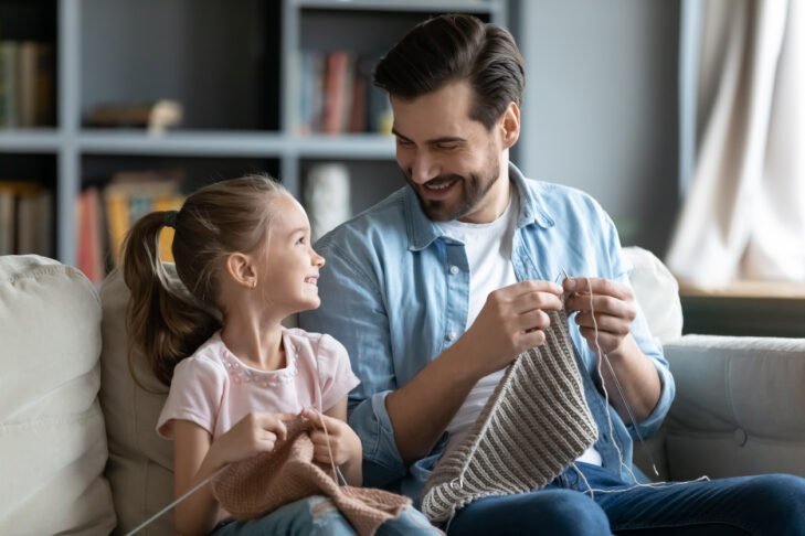 Smiling young father and little preschooler daughter sit on couch in living room knit with needles together, happy dad and small girl child have fun involved in favorite hobby family activity at home