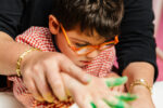 Special education, needs, parent or teacher with child or student a disabled boy is painting his birthday wreath with his hands with the help of the teacher. you see the table, the brush, acrylic paint colors green, yellow, red.plastic art exercises.