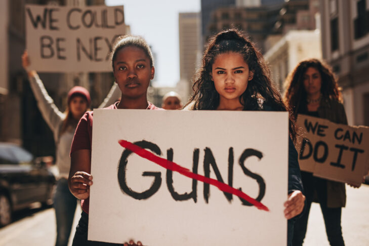 Two girls holding a banner with word guns strikethrough. Women holding sign that says not guns at a rally.