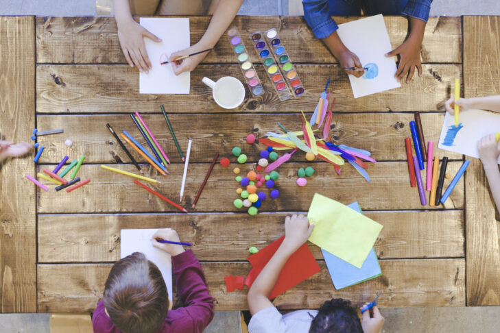 Aerial overhead view of a multi-ethnic group of elementary age children doing arts and crafts. The kids are seated around a large table that is covered by coloring and painting supplies. The creative kids are having fun and sharing.