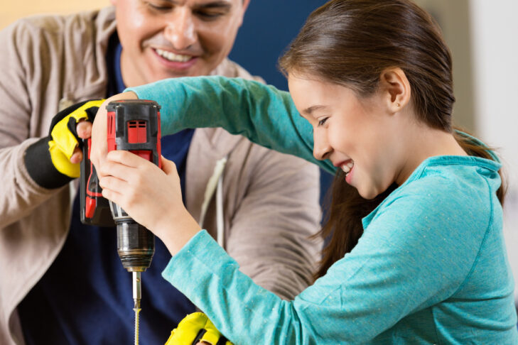 Mid adult Hispanic father teaches daughter how to use power drill in workshop. The elementary age girl smiles confidently as she uses the drill. The man is wearing fingerless gloves