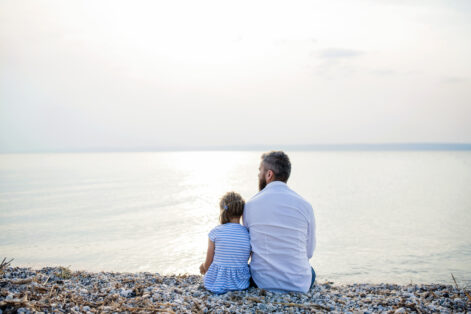 Rear view of father and small daughter on a holiday sitting by the lake or sea.