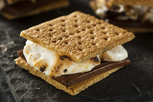 S'mores smores Close up image of S'mores dessert with roasted marshmallows and a chocolate bar sandwiched between two square graham crackers. The dessert is displayed on a dark gray background, with two more S'mores in the background. The background is slightly out of focus.