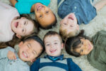 A multi-ethnic group group of young children lay down in a circle on a living room carpet with their heads together all brightly smiling at the camera.