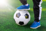 Children's foot of the winner in sports shoes sneaker stands on a soccer ball against a background of grass. Close-up street shot of game and training with victory.