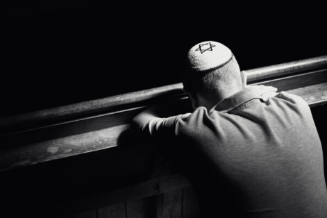 Monochrome image depicting a young Jewish adult man in his 30s inside a synagogue. He has his head bowed in prayer and he is wearing the traditional Jewish skull cap - otherwise known as a kippah or yarmulke - on his head. The man has a beard and the background of the synagogue is blurred out of focus. Horizontal color image with copy space.