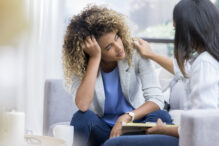 A young woman sits on a couch with her unrecognizable therapist.  She puts her head in her hand as she looks out the window with a sad expression.  Her therapist puts a hand on her shoulder.