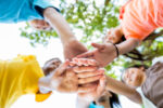 Diverse active school kids have their hands together in a circle. They are playing together at the park or school or they are at a sporting event. They are wearing bright colors. Photo shot from low angle viewpoint looking up at their hands. Focus is on the hands. Their faces and trees are blurred. It is a bright sunny day.