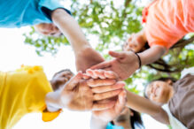 Diverse active school kids have their hands together in a circle. They are playing together at the park or school or they are at a sporting event. They are wearing bright colors. Photo shot from low angle viewpoint looking up at their hands. Focus is on the hands. Their faces and trees are blurred. It is a bright sunny day.