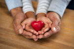 Family holding small red heart in hands on wooden background. Top view of father and daughter hands protect heart. High angle view of indian man and little girl hands holding red heart: adoption foster family, hope, gratitude and insurance concept