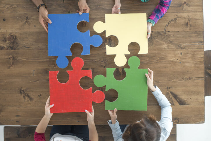 A multi-ethnic group of young adults are putting four puzzle pieces together on a table. The high angle view showing the red, blue, green, and yellow puzzle pieces.