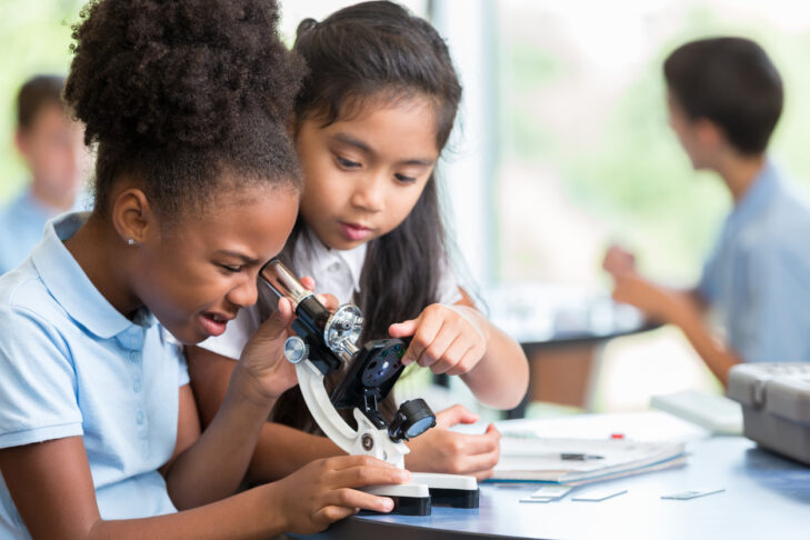 Focused African American and Asian schoolgirls help one another with their assignment in science class at a STEM elementary school. The African American girl is looking at something with a microscope.
