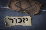 closeup of a ragged jewish badge and a yellowish piece of paper with the word yizkor, remember in hebrew and the name of a prayer in memory of deceased beloveds, on a gray rustic wooden surface