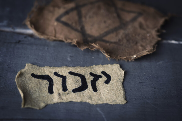 closeup of a ragged jewish badge and a yellowish piece of paper with the word yizkor, remember in hebrew and the name of a prayer in memory of deceased beloveds, on a gray rustic wooden surface