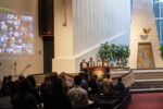 Erev RH Community Service in the Temple Israel Sanctuary with zoom participants projected on the wall, onsite participants standing at their seats and 4 clergy at the bima