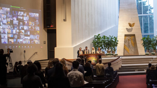 Erev RH Community Service in the Temple Israel Sanctuary with zoom participants projected on the wall, onsite participants standing at their seats and 4 clergy at the bima