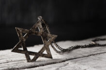 closeup of an old and rusty pendant in the shape of the star of david on a white rustic wooden surface, against a dark background