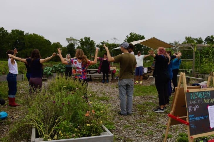 A large group of smiling people reach their arms outward, in front of raised plant beds and an easel which reads Nourish, Join Us Anytime.