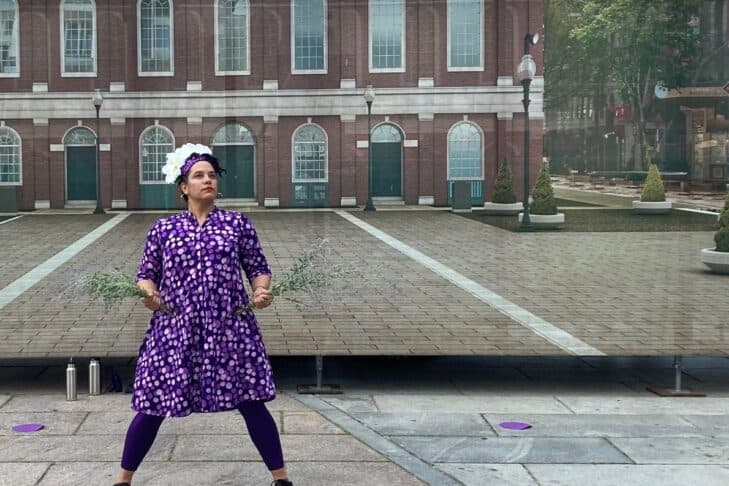 Jess, in purple and a crown, strikes a wide legged stance, hands holding fresh Russian sage in front of a trompe l’oeil image of Faneuil Hall Plaza.