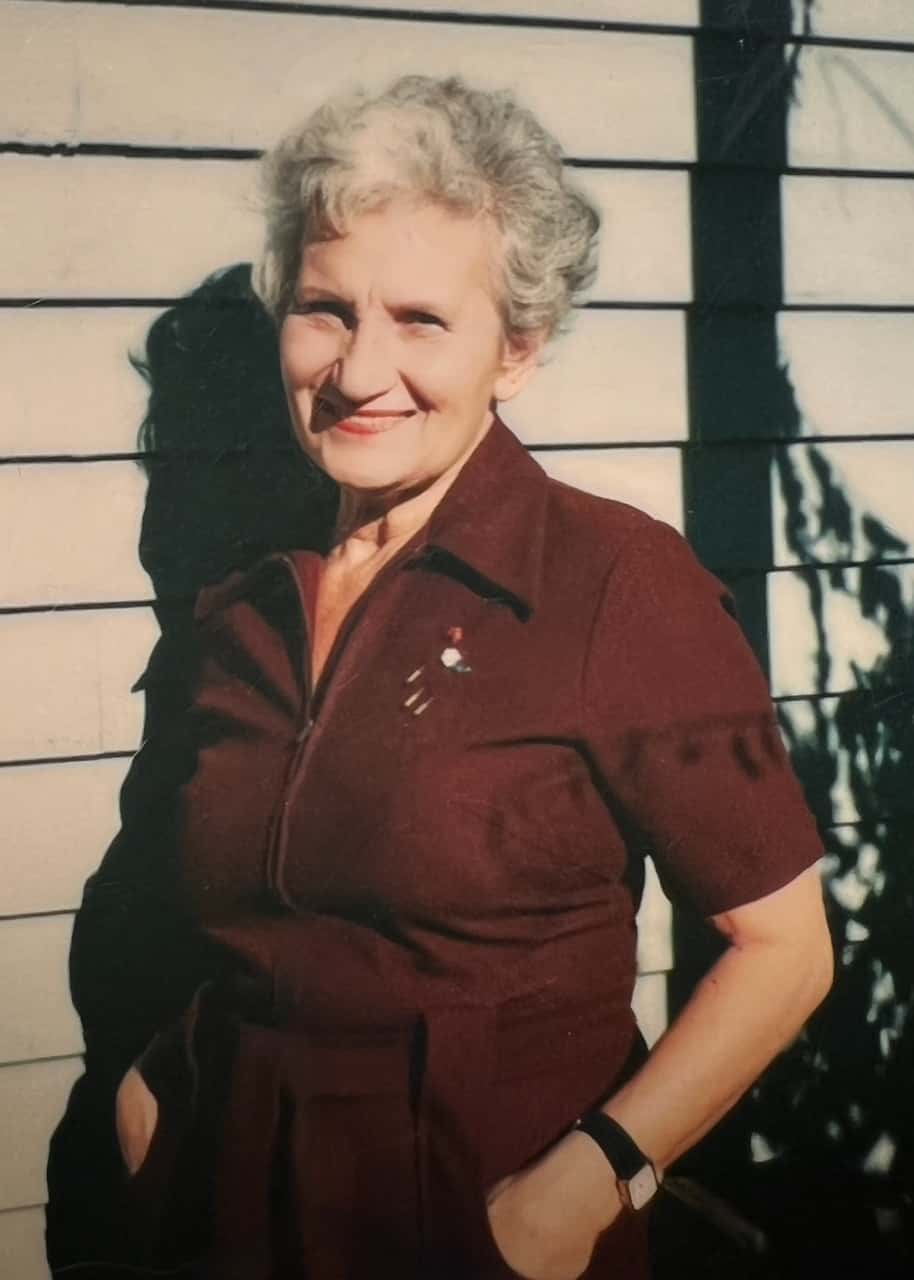 A silver haired woman in a burgundy jumpsuit smiles warmly at the camera, with red lipstick and her hands in her pockets.