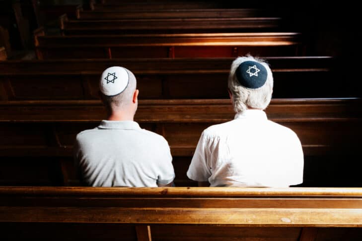 Close up image depicting a rear view of two Jewish men sitting together inside a synagogue. They have their heads bowed in prayer and they are wearing the traditional Jewish skull cap - otherwise known as a kippah or yarmulke - on their heads. Horizontal color image with copy space.