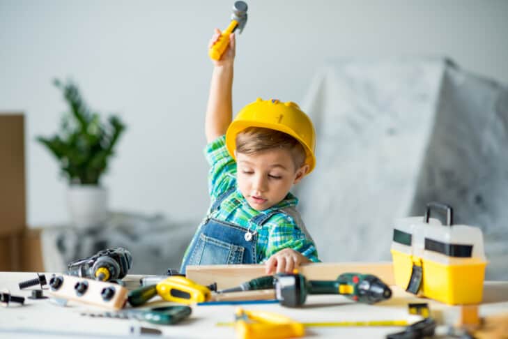 Cute little boy in yellow hard hat holding wooden plank and toy hammer