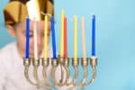 Kid celebrating Hanukkah Israel holiday. Little Jewish boy puts candles on traditional menorah with eight candles. Selective focus on yellow candle.
