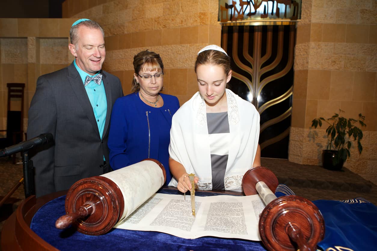A thirteen year old girl in a Jewish Temple is reading the Hebrew Torah on her Bat Mitvah with her parents at her side