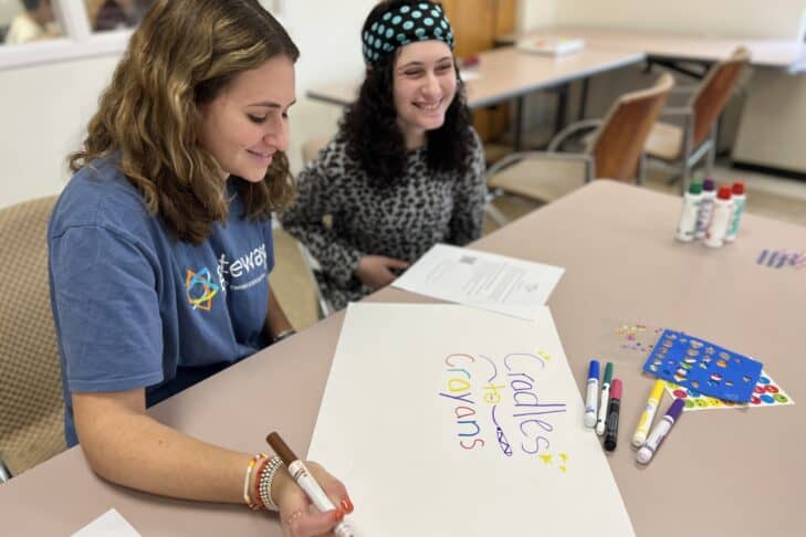 A Gateways teen volunteer writes something on a poster for Cradles to Crayons, as a student siting next to her smiles and watches