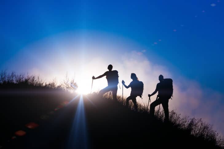 Group of people silhouettes walking toward mountain summit with backpacks and hiking trekking gear meeting uprising sun sunbeams and blue sky of background