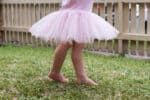 Toddler girl in pink tutu dancing on grass in front garden with picket fence (cropped)