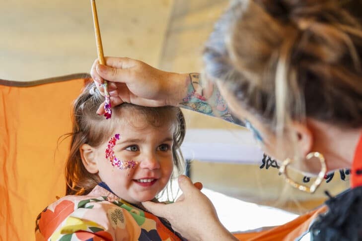A young girl getting glitter painted on her face by an unrecognizable face painter. She is sitting at a stall, part of the Whitley bay Food Festival.