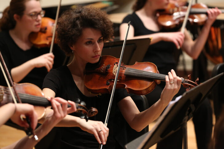 A young woman with dark curly hair wearing a black dress plays violin.