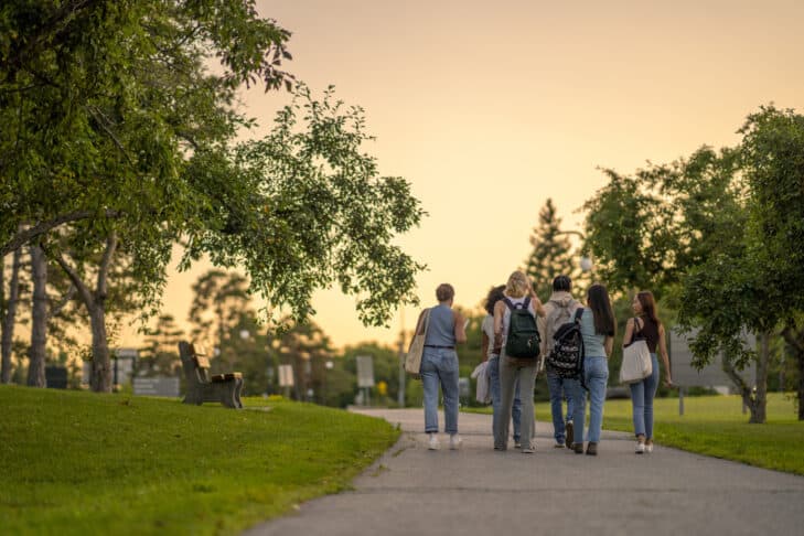 A group of University students are seen from behind walking outside on campus as they make their way to class.  They are dressed casually and have backpacks on as they talk together and make their way to school for the day.