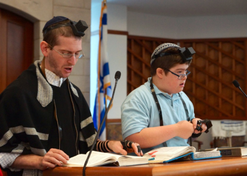 A student with down syndrome and a teacher stand next to each other on the bimah wrapping tefillin