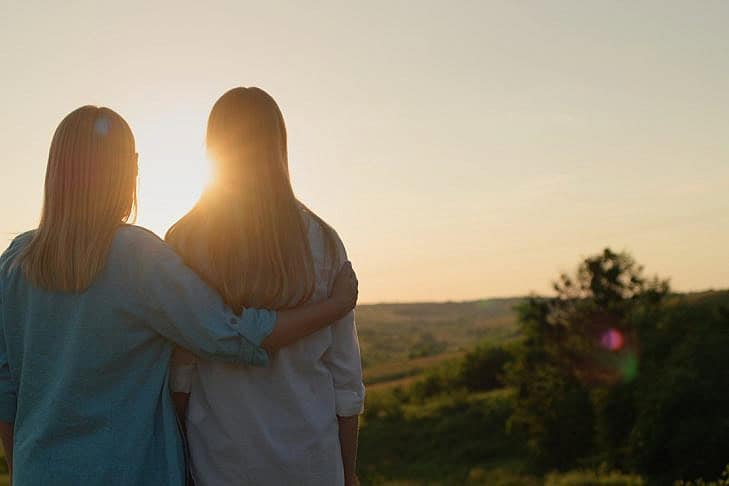 A woman hugs her teenage daughter and together they watch the sun go down over a picturesque valley A woman hugs her teenage daughter and together they watch the sun go down over a picturesque valley. Back view.