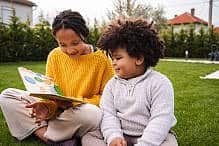Older sister reading a book to younger brother outdoors Children enjoying storytelling while sitting together on green grass in a backyard