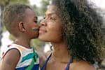 Little black 4 years old boy who painted his face with LGBTQ flags, kissing his mother's cheek at city street during marriage equality pride celebration. LGBTQ African American mother hugging her child boy during enjoying street carnival LGBTQ parade. Little black 4 years old boy who painted his face with LGBTQ flags, kissing his mother's cheek at city street during marriage equality pride celebration. LGBTQ African American mother with afro hair style hugging her child boy during enjoying street carnival LGBTQ parade.