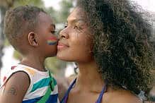 Little black 4 years old boy who painted his face with LGBTQ flags, kissing his mother's cheek at city street during marriage equality pride celebration. LGBTQ African American mother hugging her child boy during enjoying street carnival LGBTQ parade. Little black 4 years old boy who painted his face with LGBTQ flags, kissing his mother's cheek at city street during marriage equality pride celebration. LGBTQ African American mother with afro hair style hugging her child boy during enjoying street carnival LGBTQ parade.