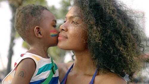 Little black 4 years old boy who painted his face with LGBTQ flags, kissing his mother's cheek at city street during marriage equality pride celebration. LGBTQ African American mother hugging her child boy during enjoying street carnival LGBTQ parade. Little black 4 years old boy who painted his face with LGBTQ flags, kissing his mother's cheek at city street during marriage equality pride celebration. LGBTQ African American mother with afro hair style hugging her child boy during enjoying street carnival LGBTQ parade.